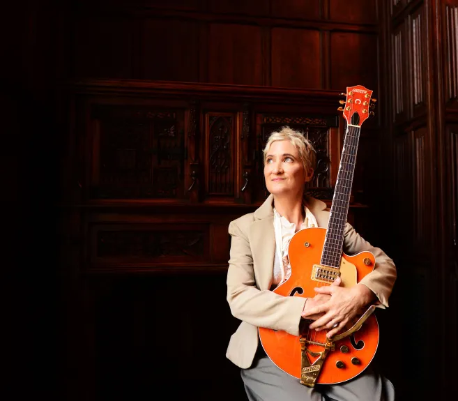 Singer-songwriter Jill Sobule sits with her orange Gretsch guitar, gazing upward in a wood-paneled room.